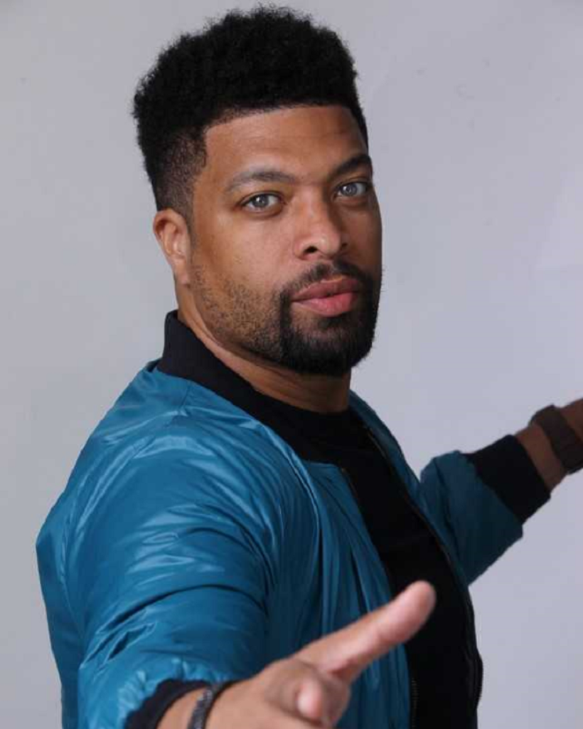 A man with a beard is wearing a blue jacket and pointing at the camera.