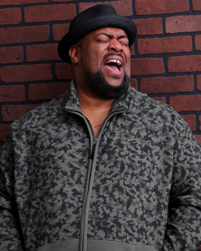 A man wearing a hat and a jacket is standing in front of a brick wall.