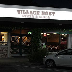 A white car is parked in front of a restaurant called village host pizza & grill.