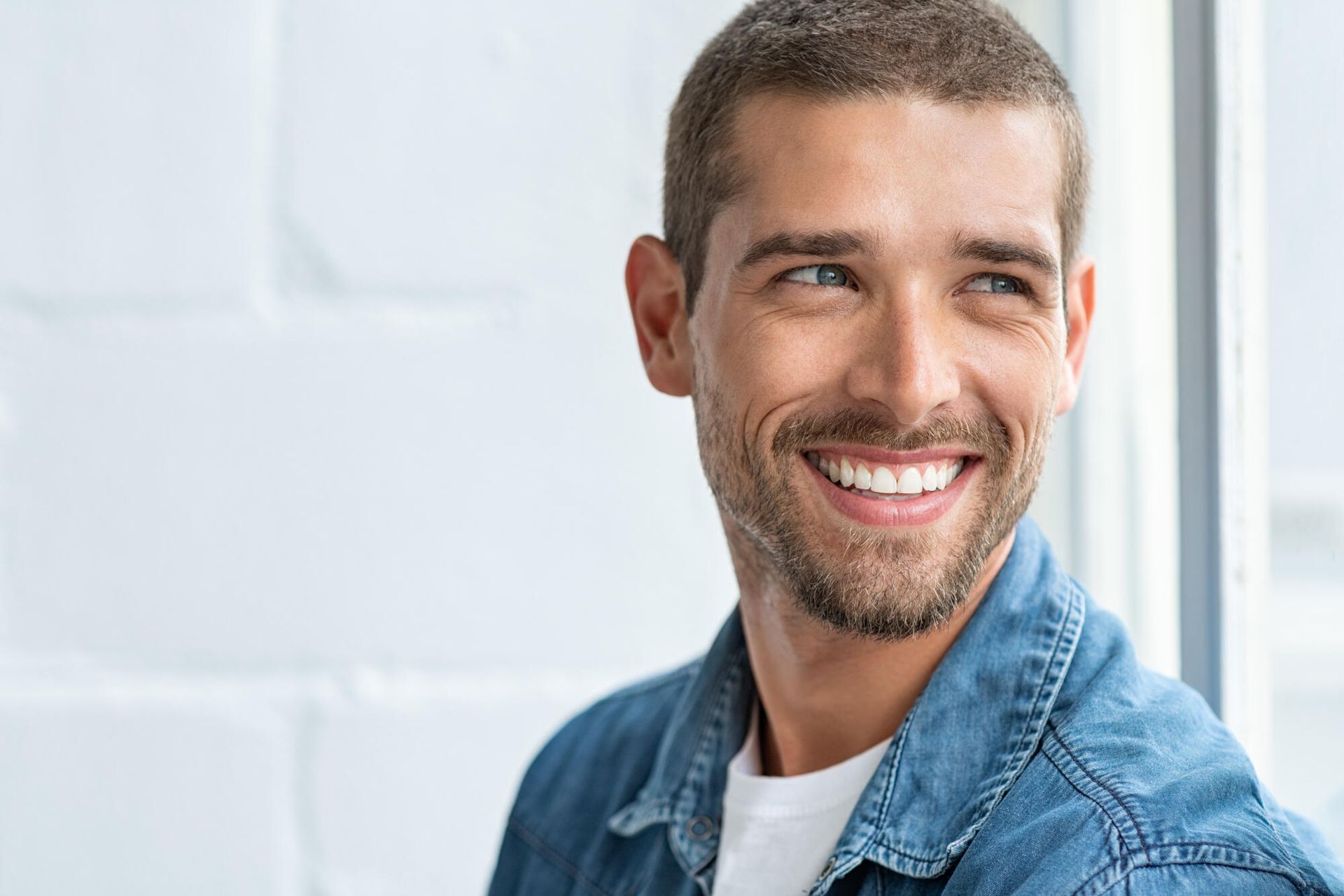 A man in a denim jacket is smiling and looking out a window.