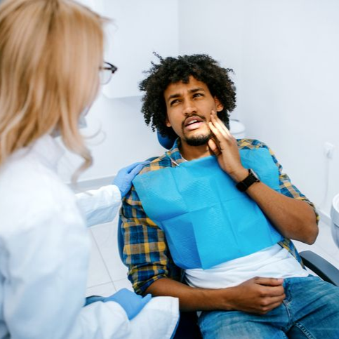 Dentist examining a patient in a blue dental bib as they hold their cheek in a clinic chair