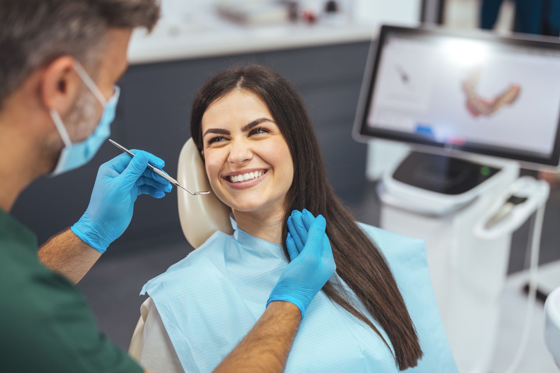 Dentist examining a patient's teeth. Smiling woman in dental chair, blue gloves, screen in background.