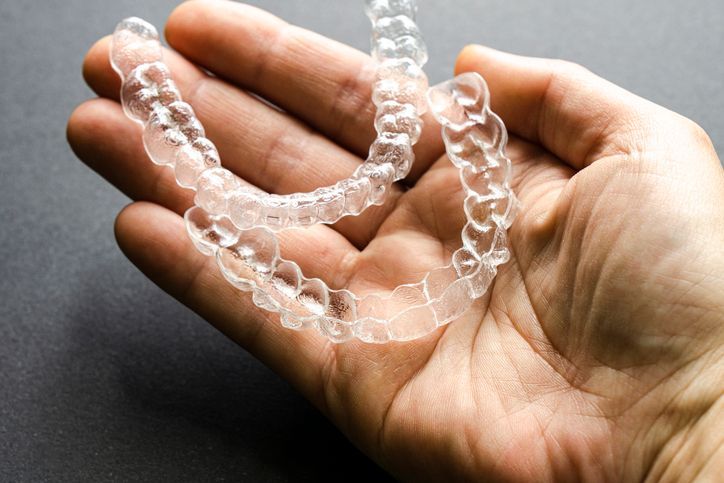 A hand holding a pair of clear plastic dental aligners against a dark gray background.