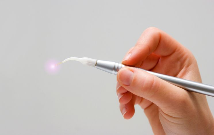 A hand holding a silver dental tool with a glowing purple tip against a plain, light gray background.