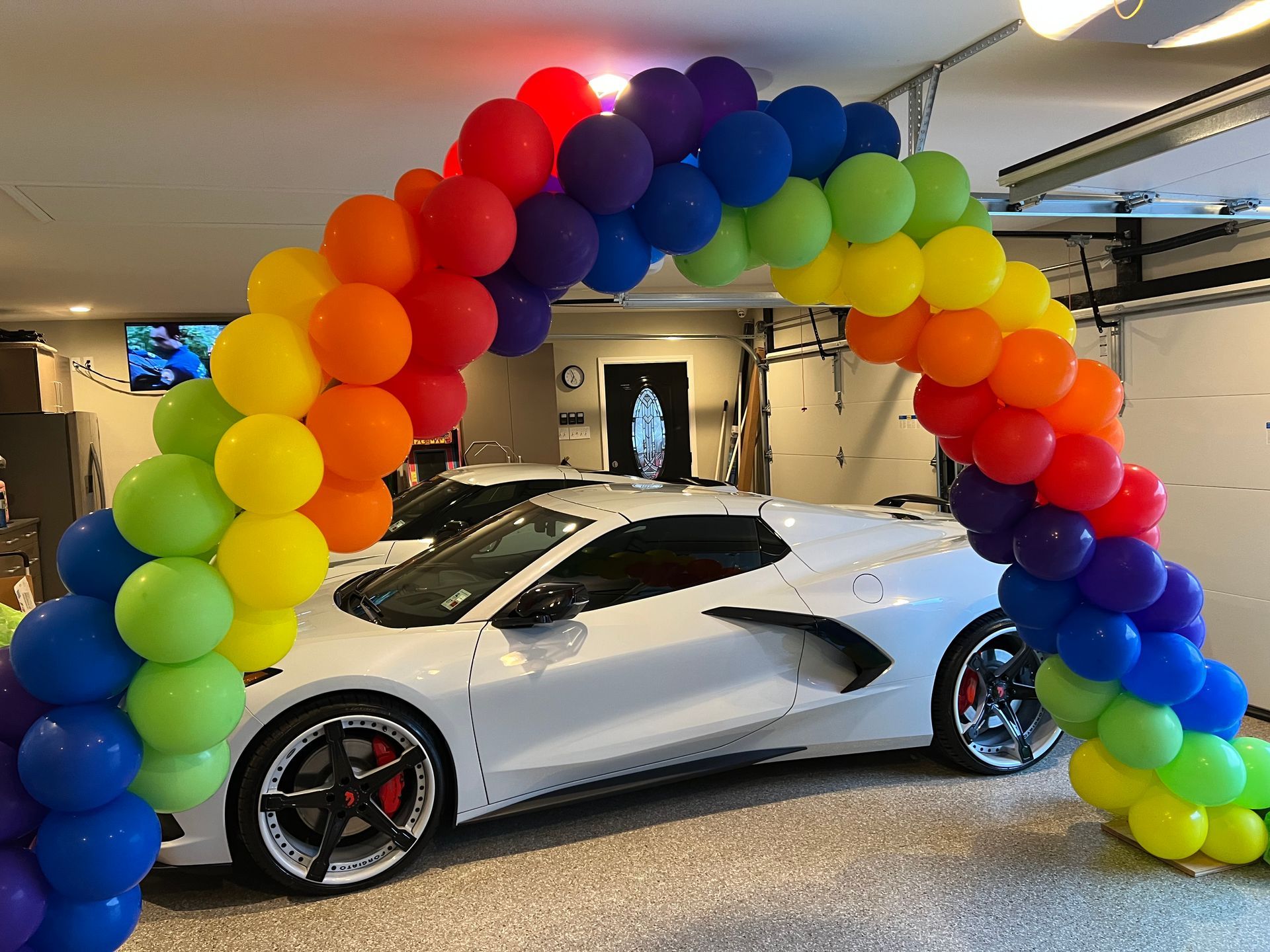 A white sports car is parked under a rainbow of balloons in a garage.