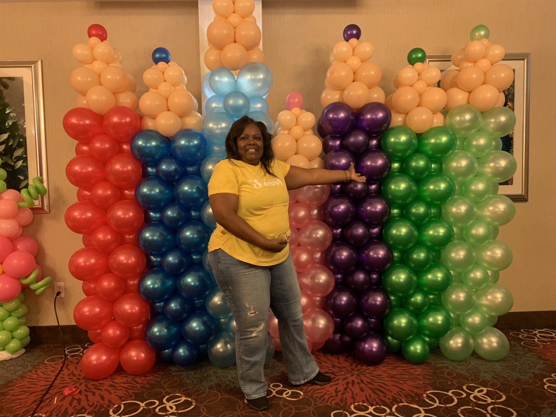 A woman in a yellow shirt is standing in front of a wall of balloons.