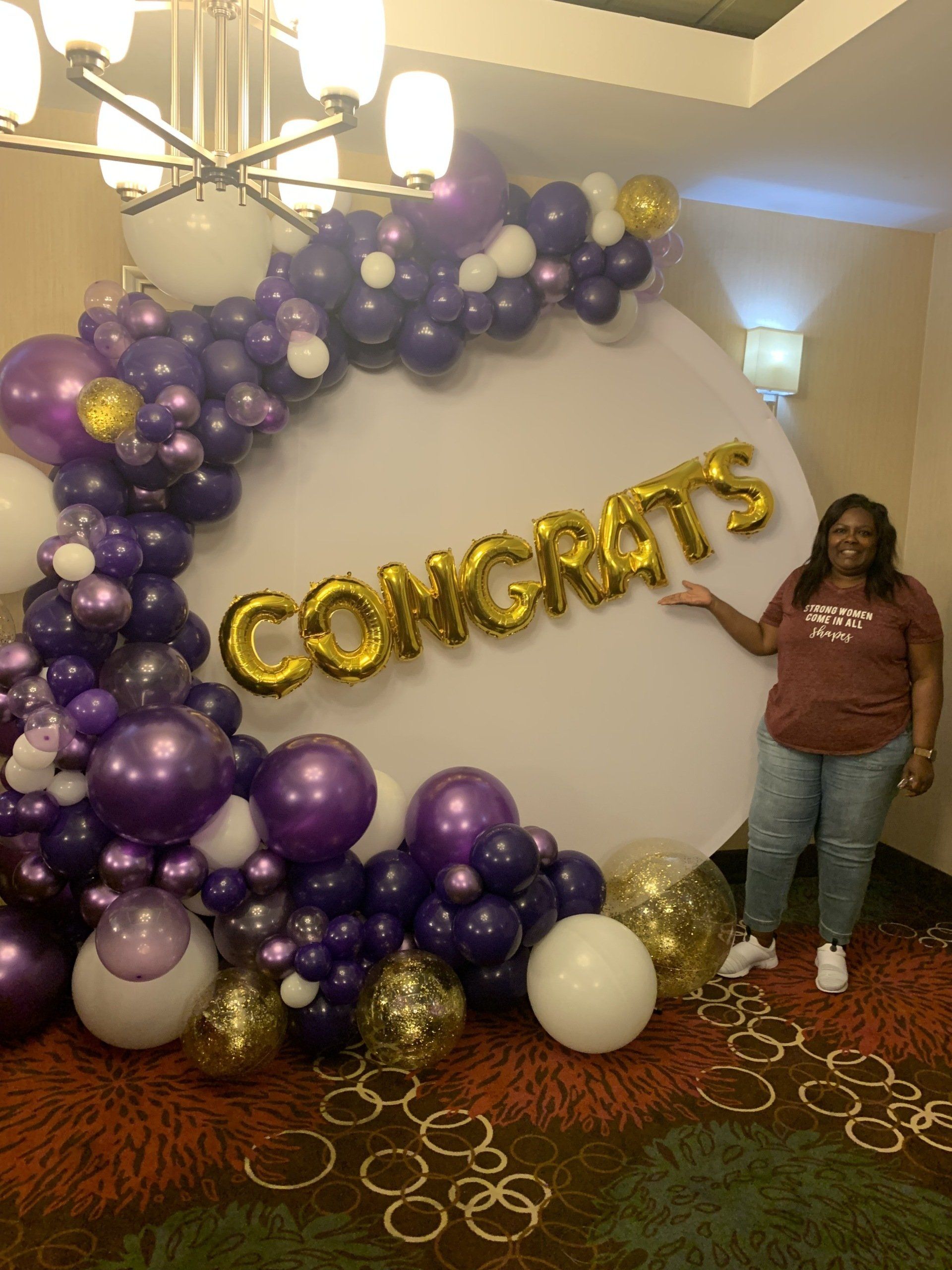 A woman is standing in front of a wall decorated with purple and gold balloons.
