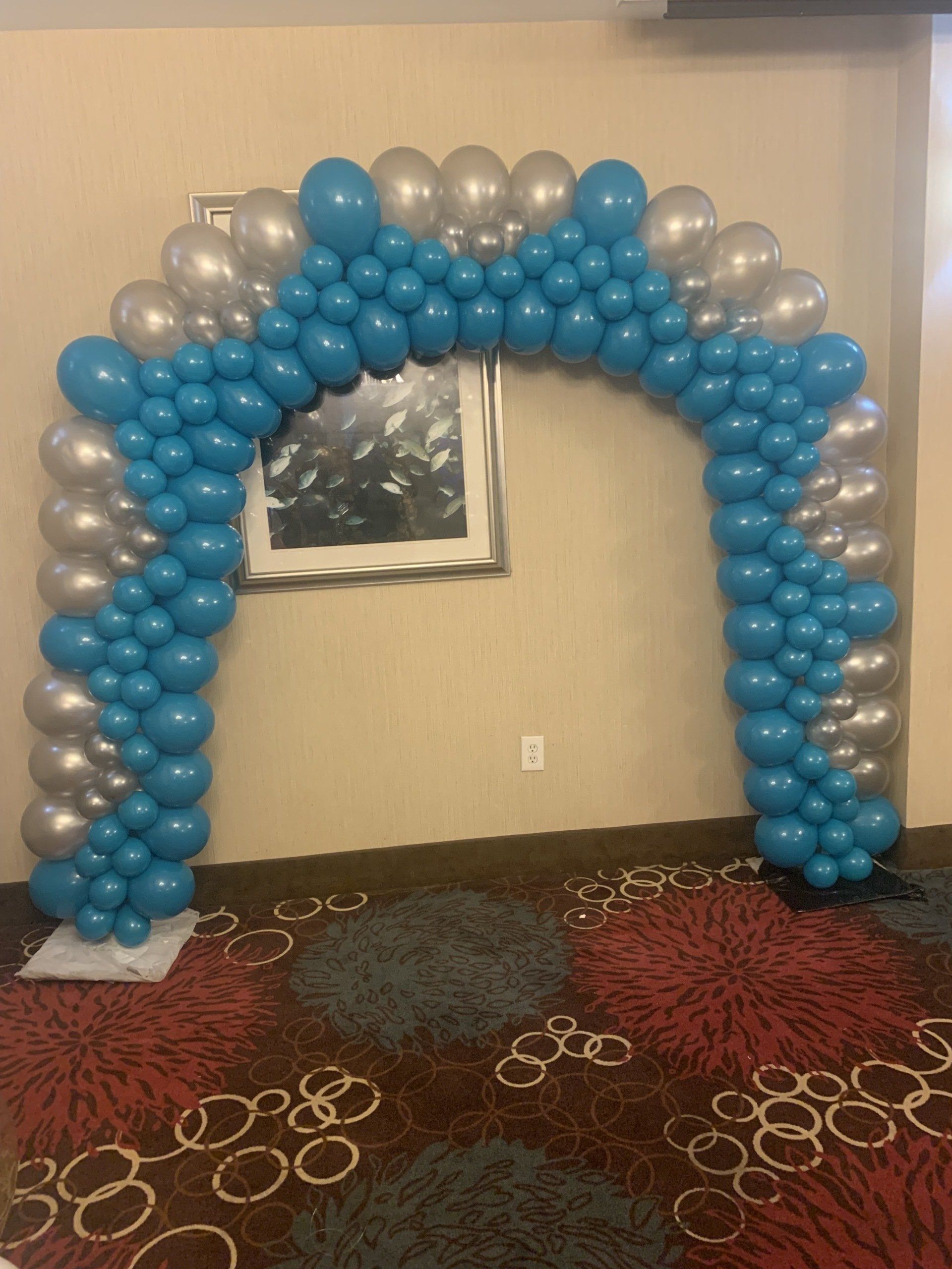 A blue and silver balloon arch in a room with a picture on the wall.