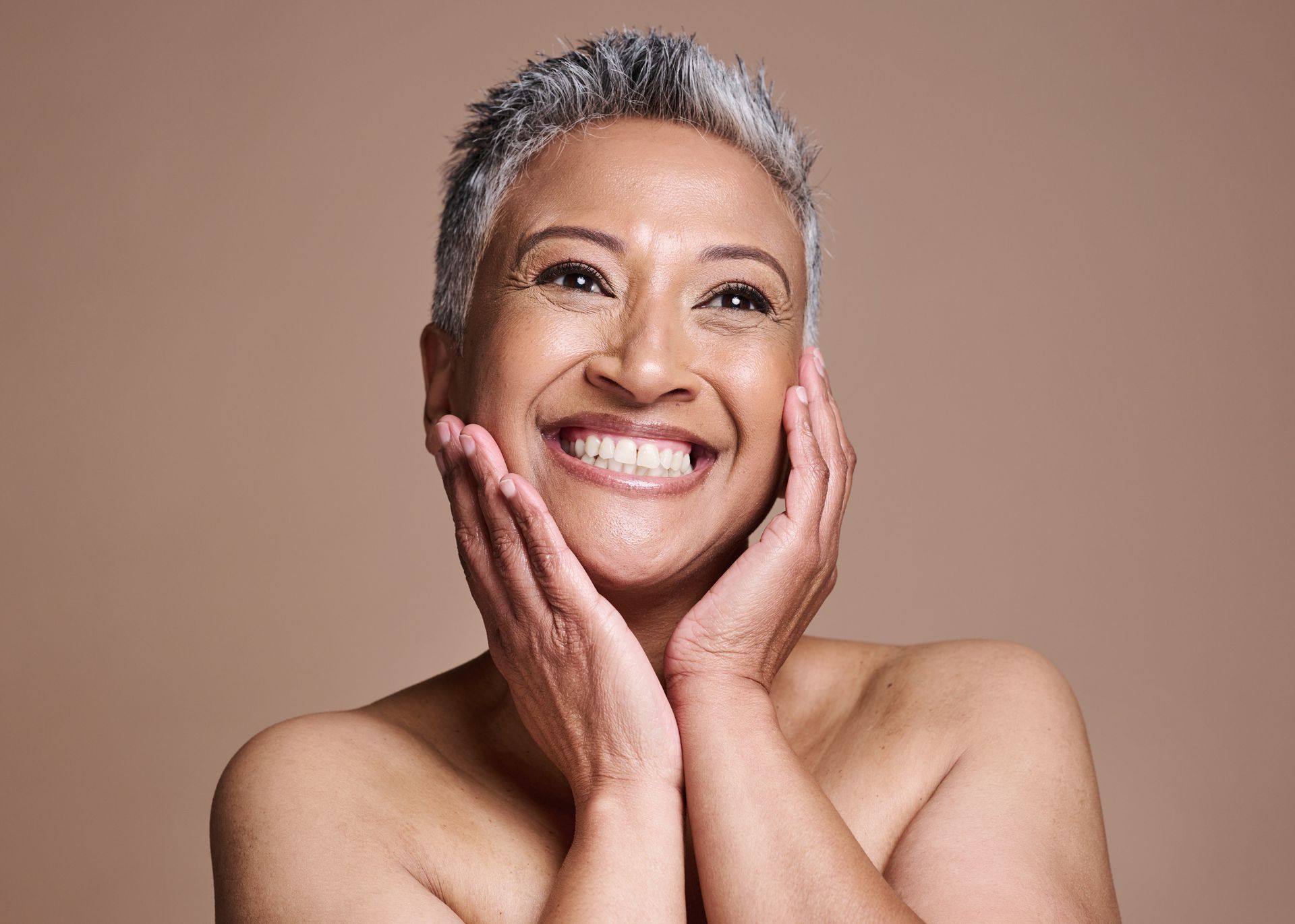 A woman with gray hair is smiling and touching her face with her hands.