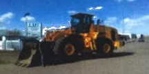 Yellow loader on pavement in front of a blue sky and a building.