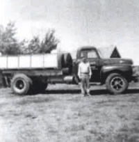 Man stands by a large, dark truck with a flatbed in a field. Black and white photo.