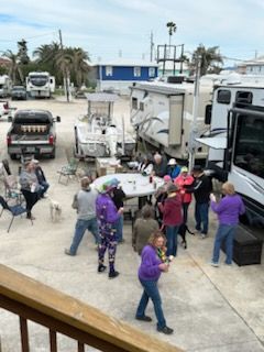 Group of people gathered around a table outdoors next to RVs; some are holding drinks. Cloudy day.