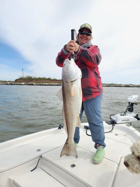 Person holding a large redfish on a boat; cloudy sky, water, and shoreline in the background.