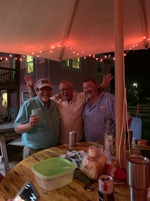 Three men smiling, arms raised, gathered around a wooden table under string lights at night.