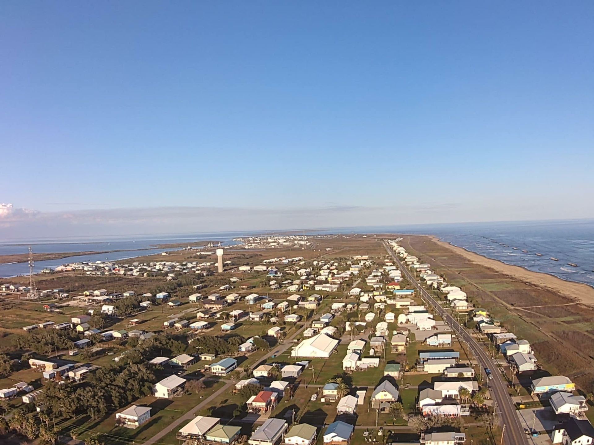 Aerial view of a coastal town with a long, narrow spit of land, buildings, and ocean under a blue sky.