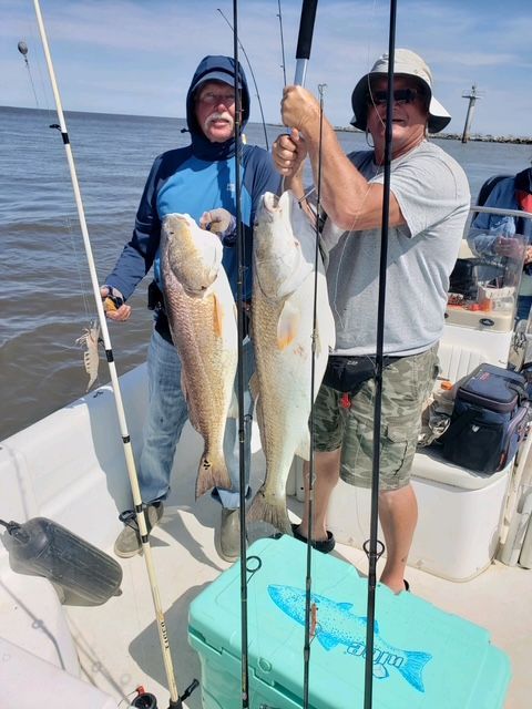 Two men on a boat hold up two redfish they caught. Light blue water, sunny day.