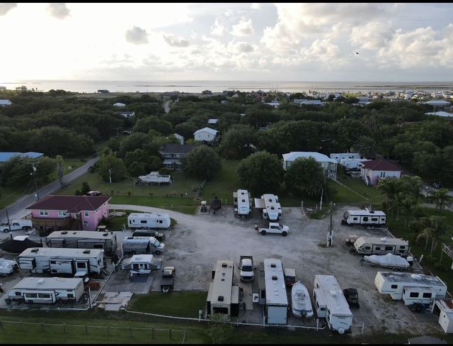Aerial view of RV park with white RVs, trees, and buildings, near water under a cloudy sky.
