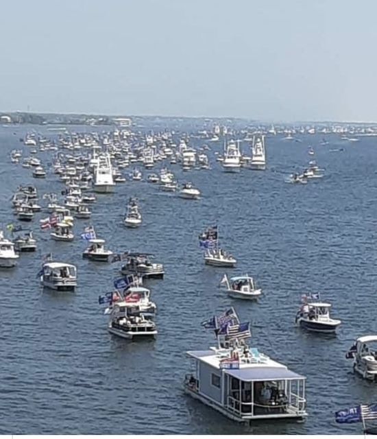 Boats of various sizes on a body of water, some with flags, a distant shoreline.