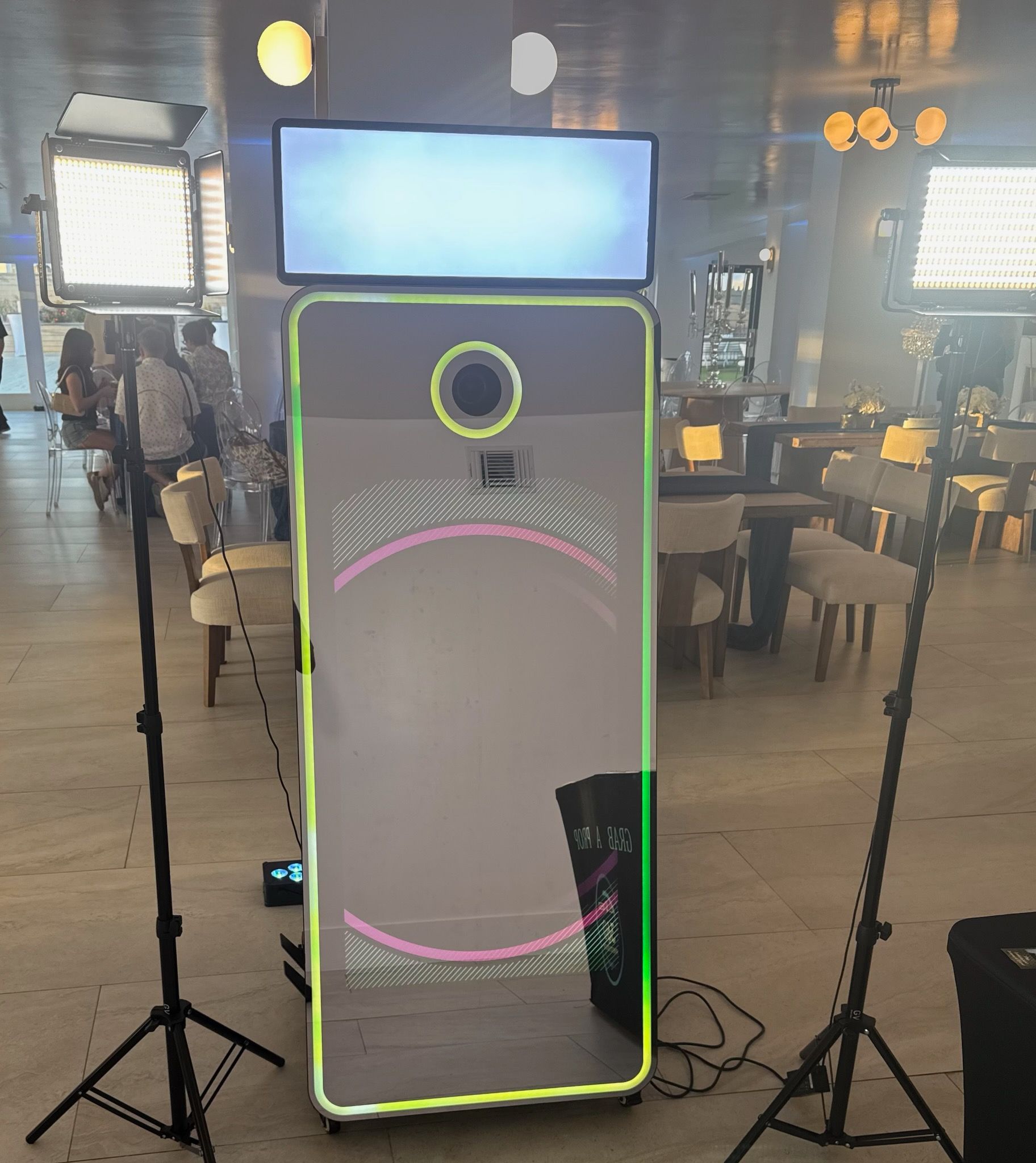 Photo booth setup at an event with a large mirror screen, lights, and a monitor. People and chairs are visible in the background.