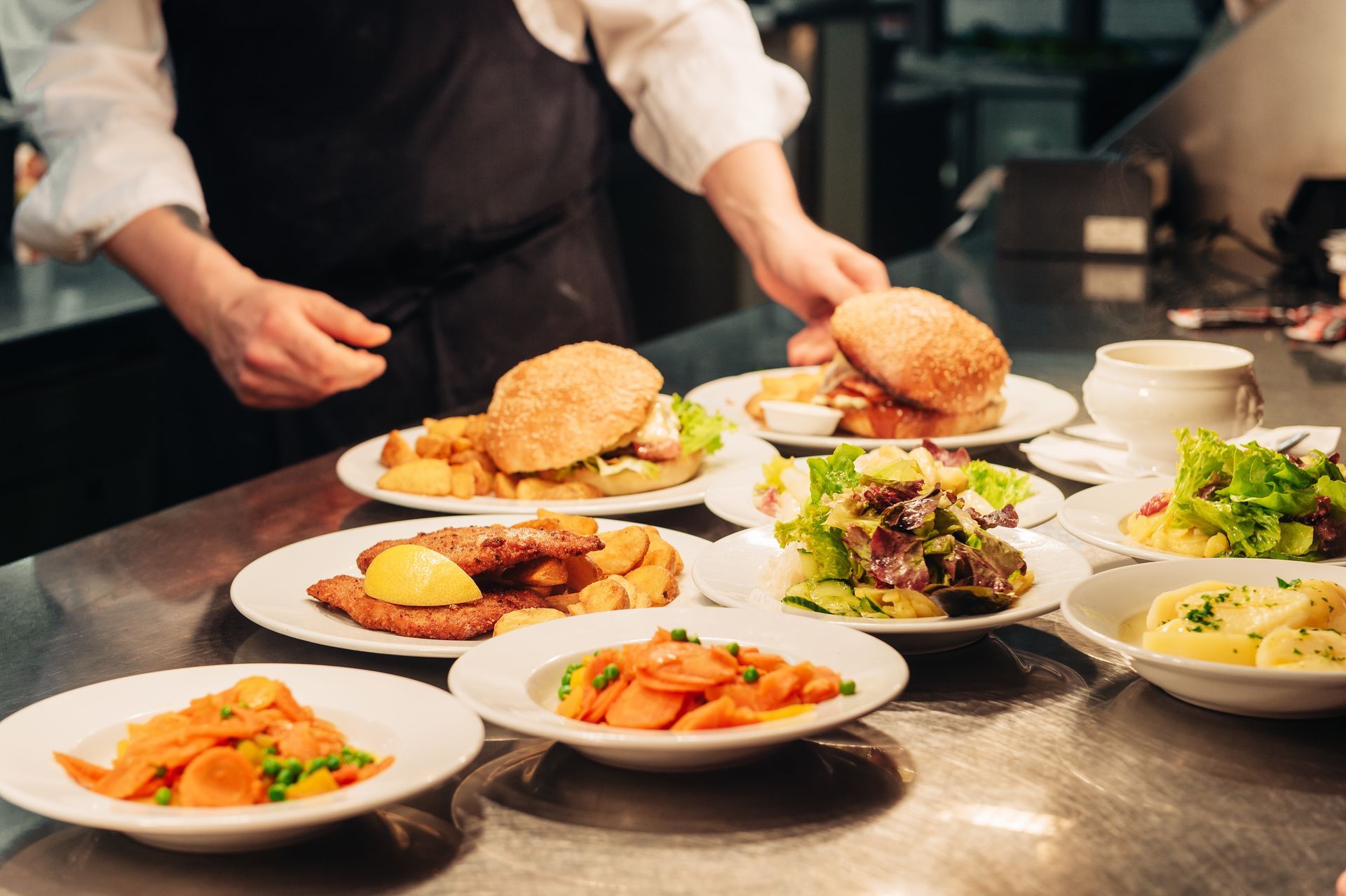 Chef placing plates of food on a stainless steel counter. Dishes include burgers, salad, and sides.