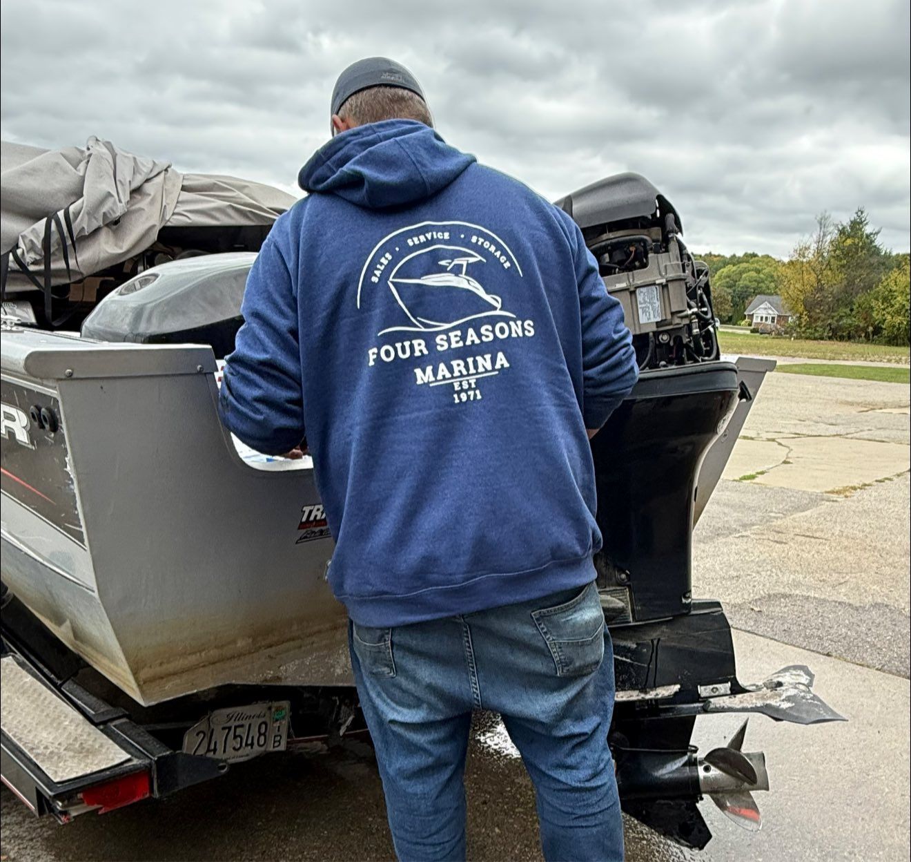 Person wearing blue hoodie, standing by a boat with motor, outdoors. Hoodie reads 