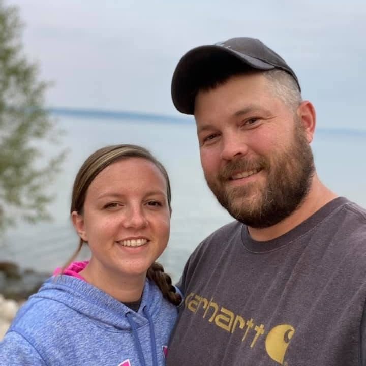 Couple smiling, posing near water. Man wearing a hat and Carhartt shirt. Woman in a hoodie.