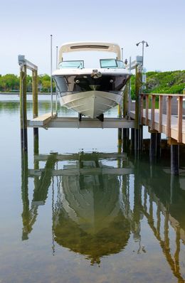 Boat on a lift, reflected in calm water.  White boat, wooden dock, blue sky, and green foliage.