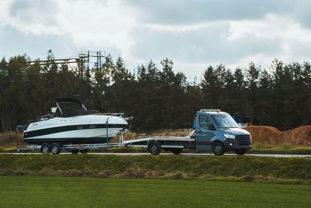 A blue flatbed truck towing a white boat on a trailer, driving on a road next to a field.
