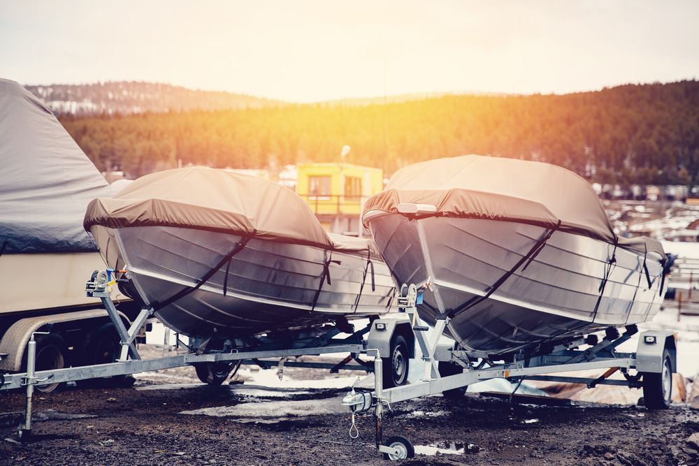 Two covered boats on a trailer, parked outside. Bright sunlight, winter setting.