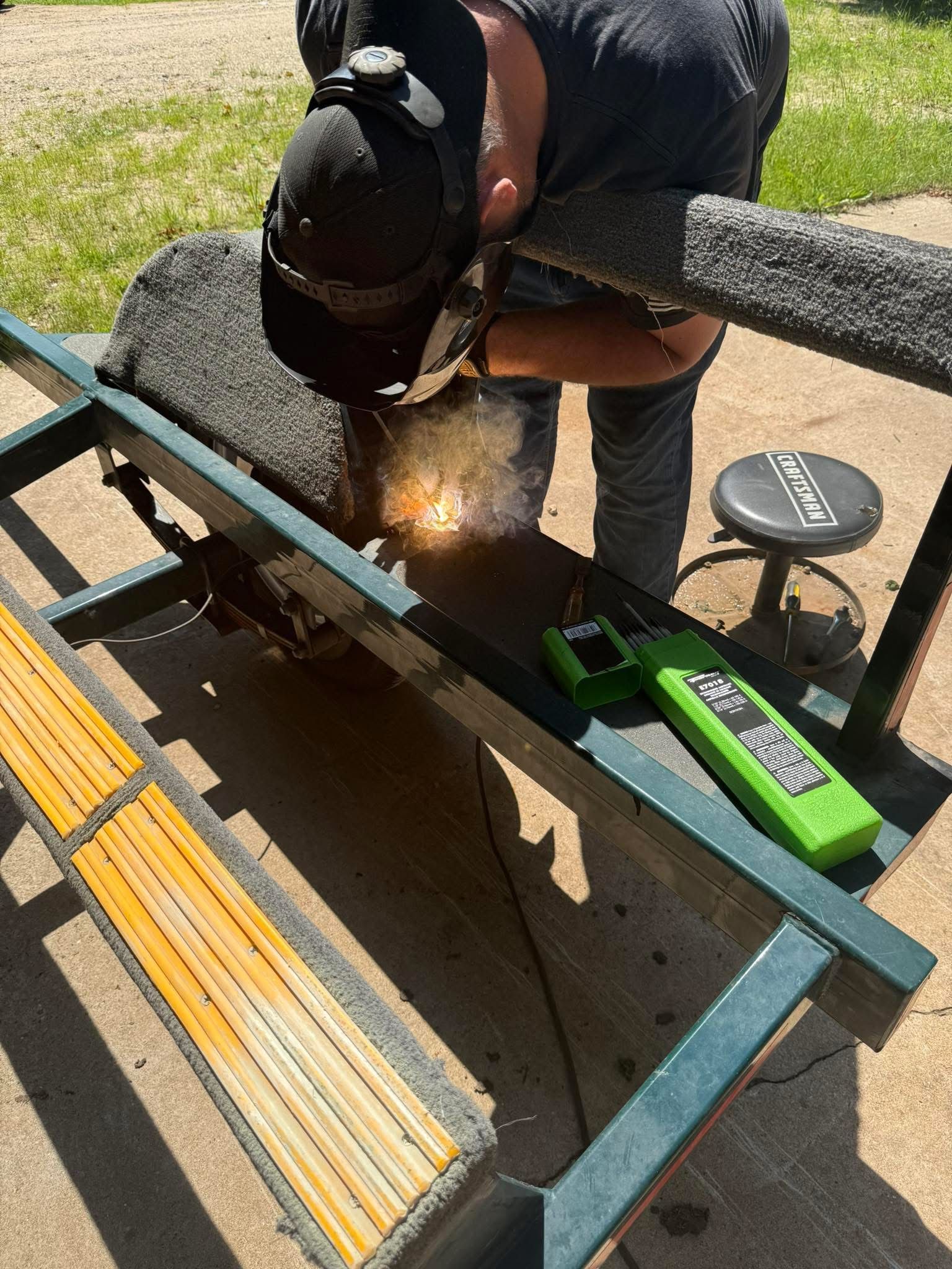 Person welding metal outdoors, sparks flying.  They wear a welding mask and hold a welding tool, positioned over metal bench.