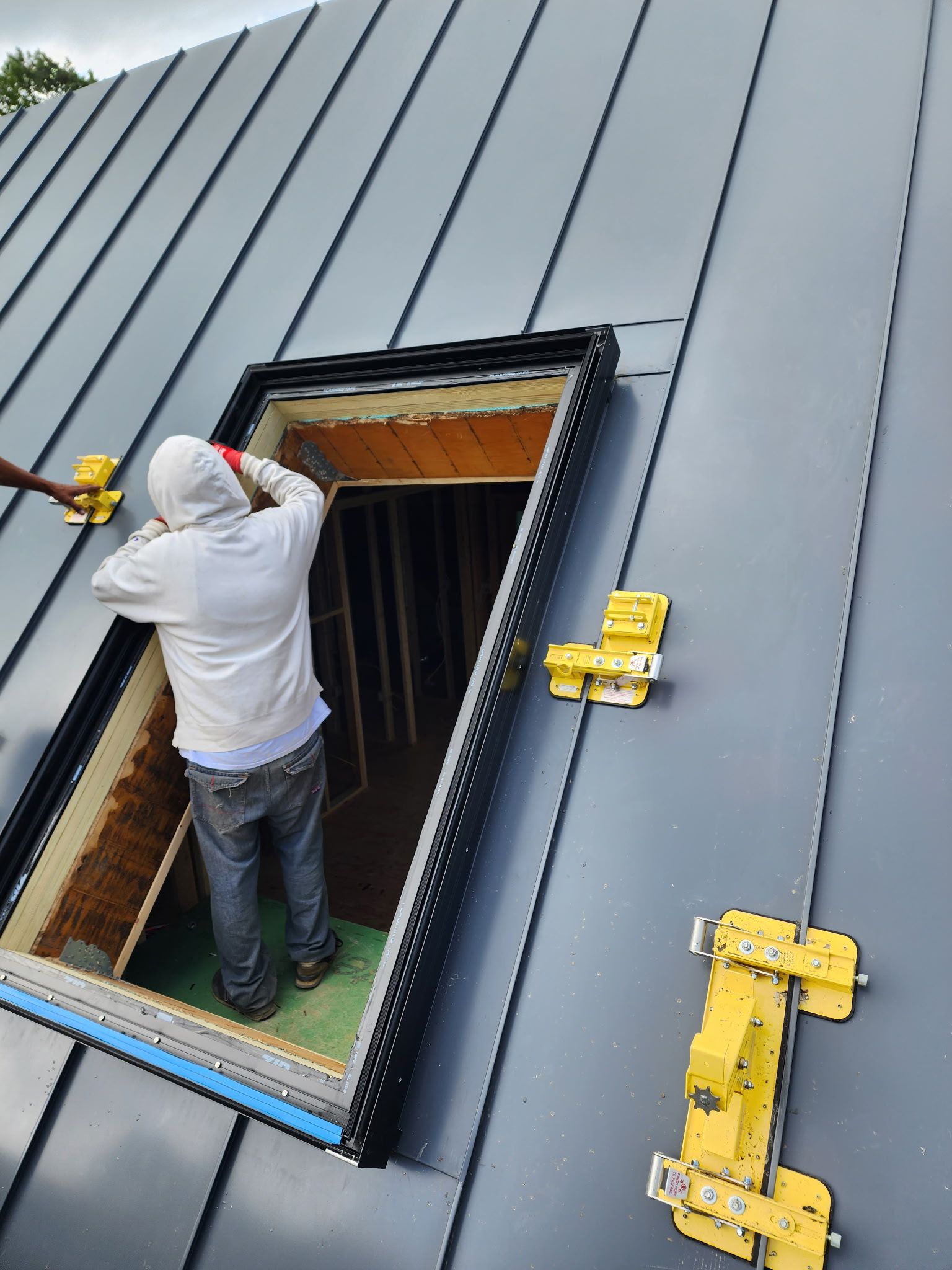 A person in a hooded sweatshirt stands inside a skylight opening on a sloped metal roof with yellow safety brackets.
