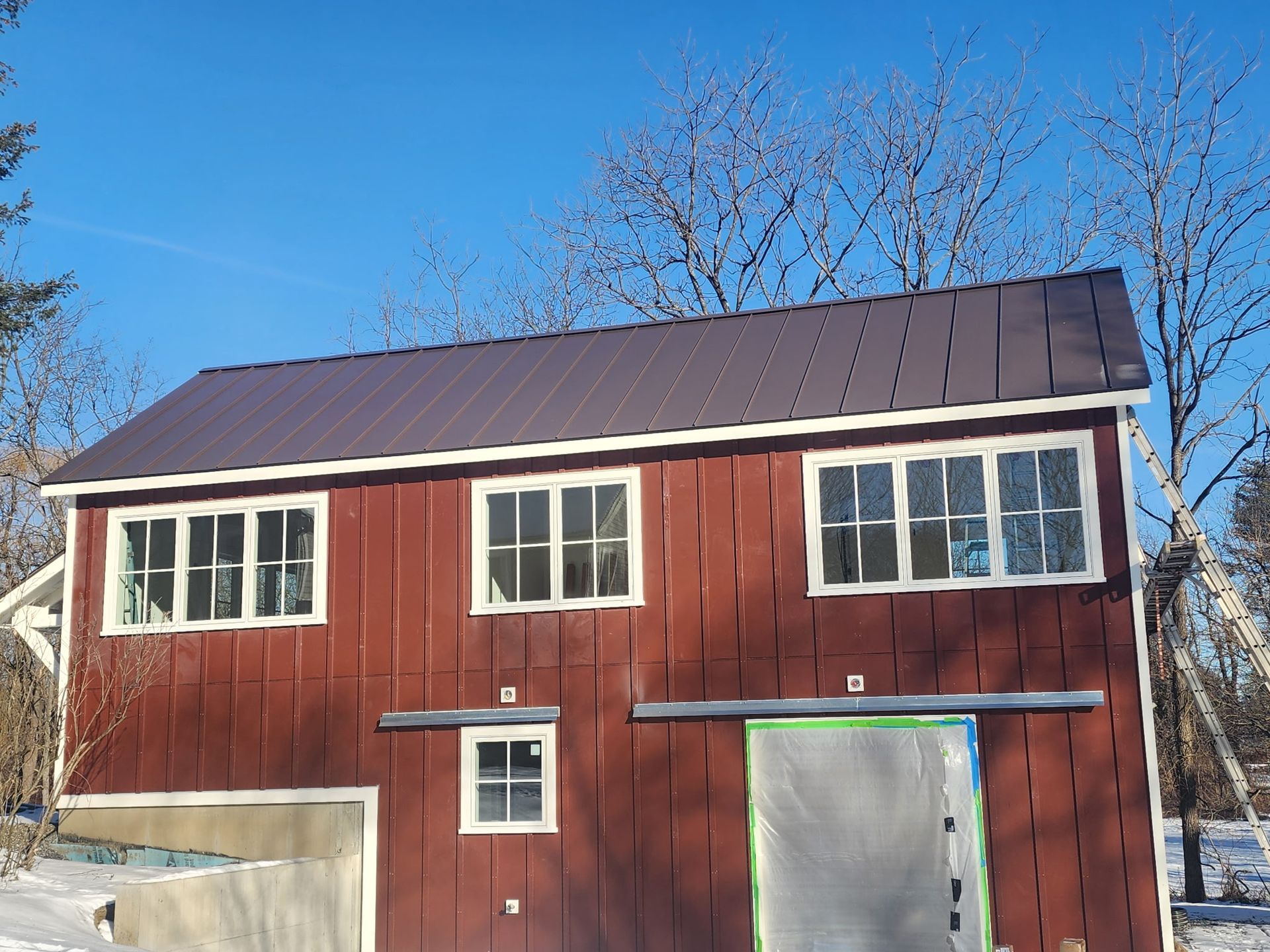 A red two-story barn with a dark metal roof, white window frames, and a partially covered ground-level door in winter.