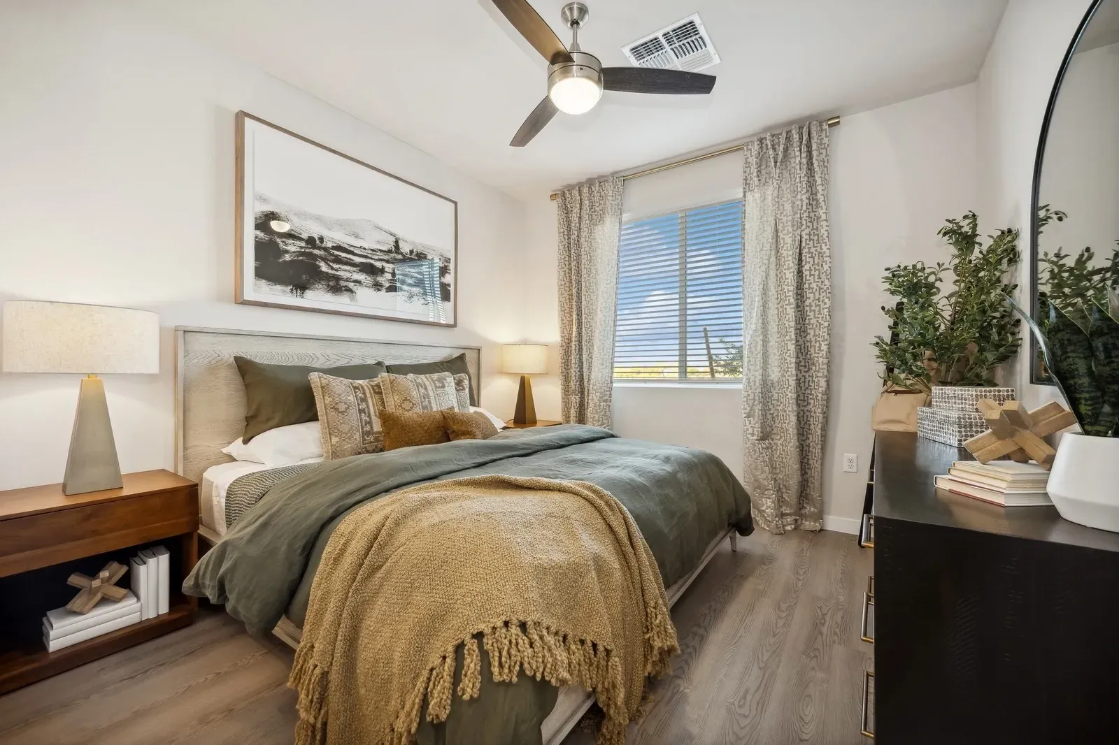 Bright bedroom with a bed, wooden nightstands, ceiling fan, and a window with patterned curtains.