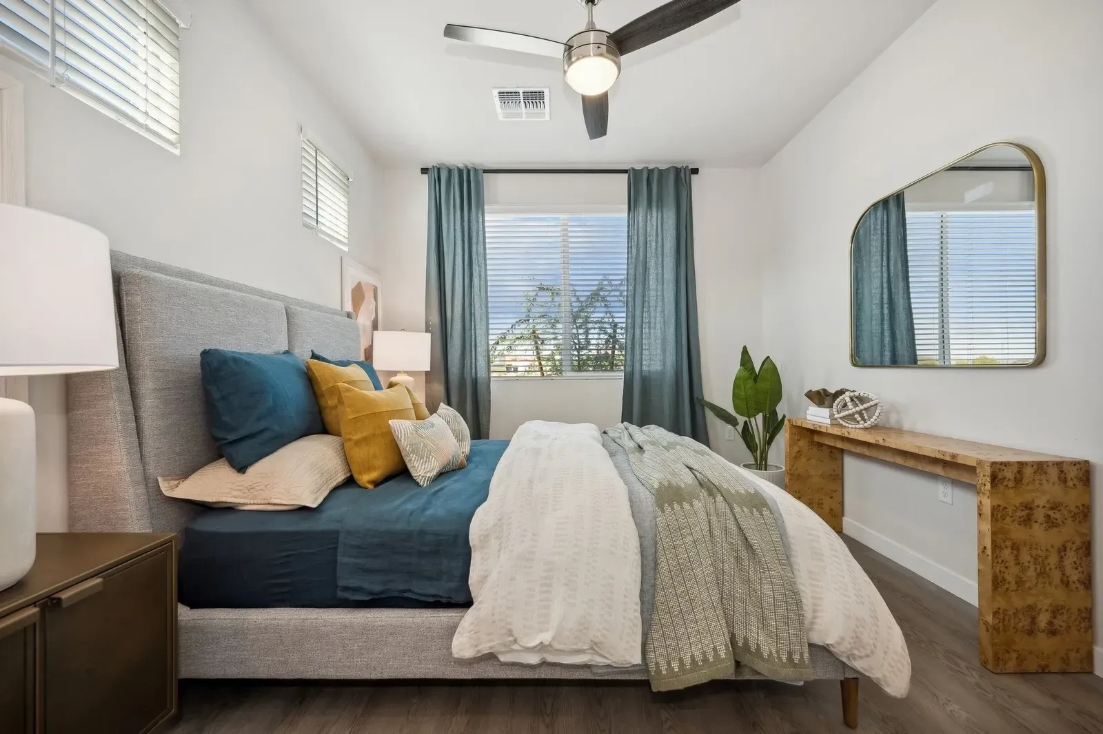 Bright bedroom with blue bedding, beige chair, window curtains, ceiling fan, and a mirrored dresser