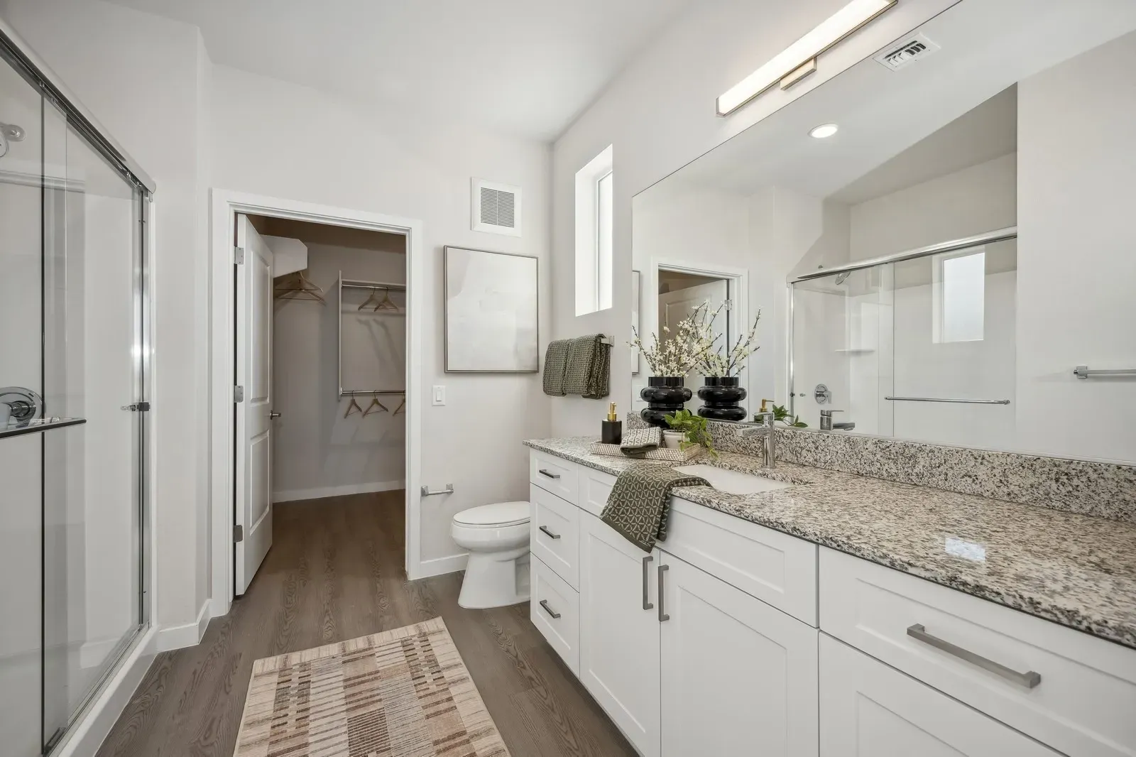 Bright modern bathroom with white vanity, granite countertop, mirror, and walk-in closet visible nearby