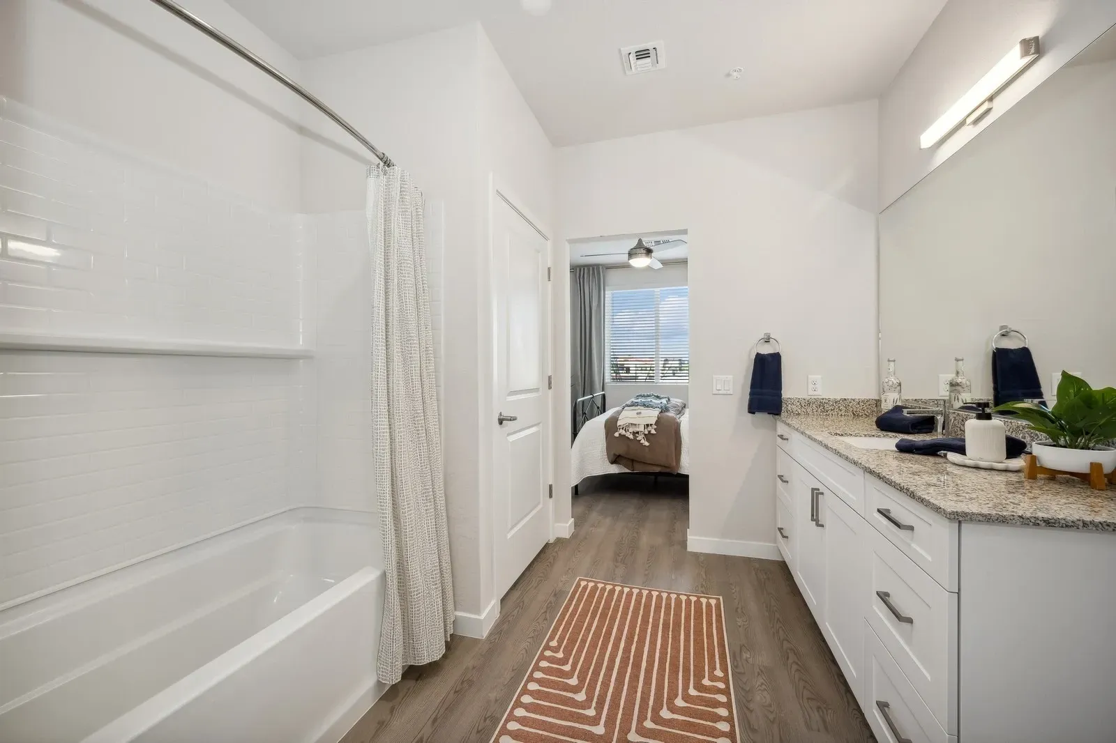 Bright bathroom with white tub, vanity, patterned rug, and doorway to a bedroom view