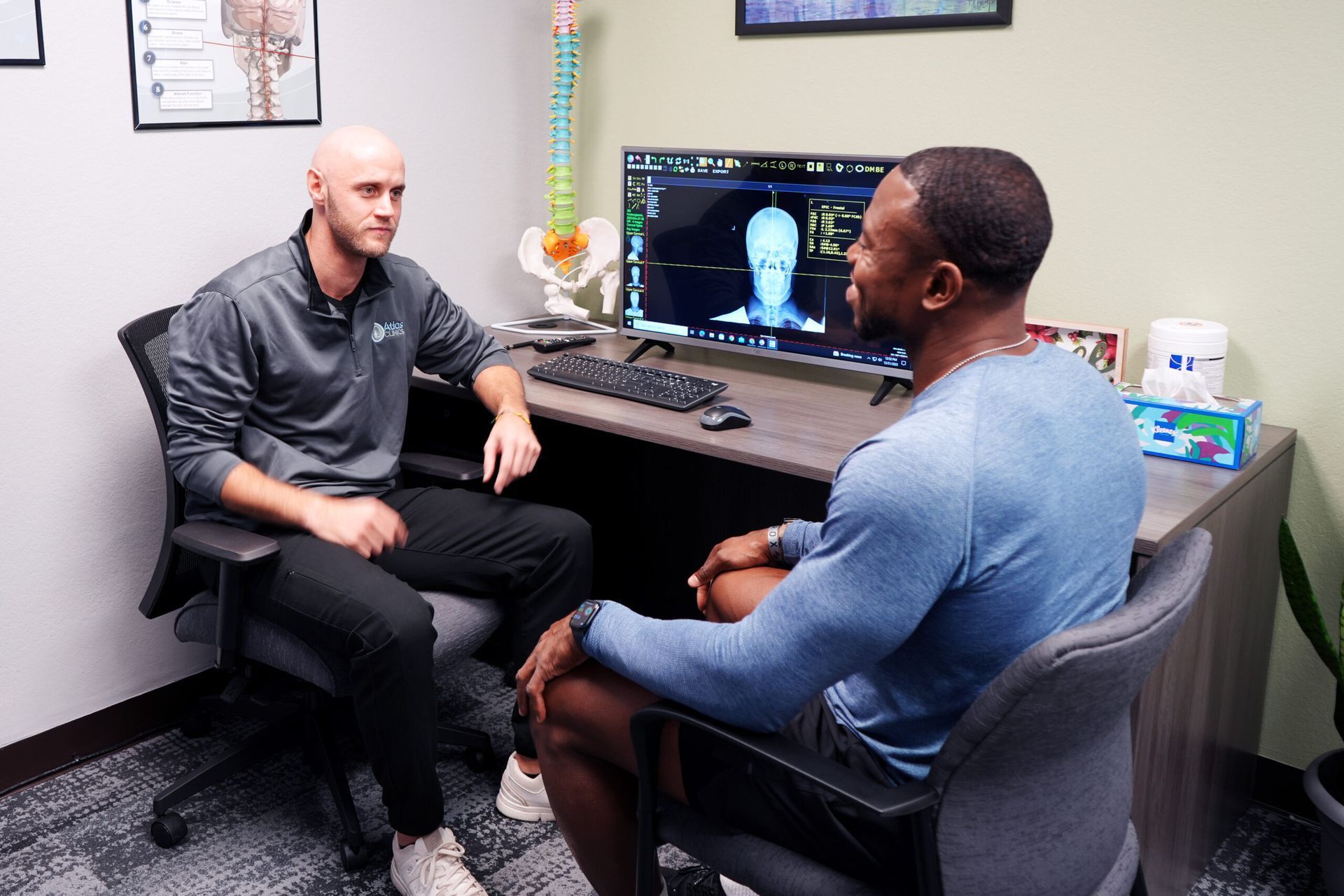 Two men seated at a desk, one speaking, the other looking at a computer screen displaying a body scan.
