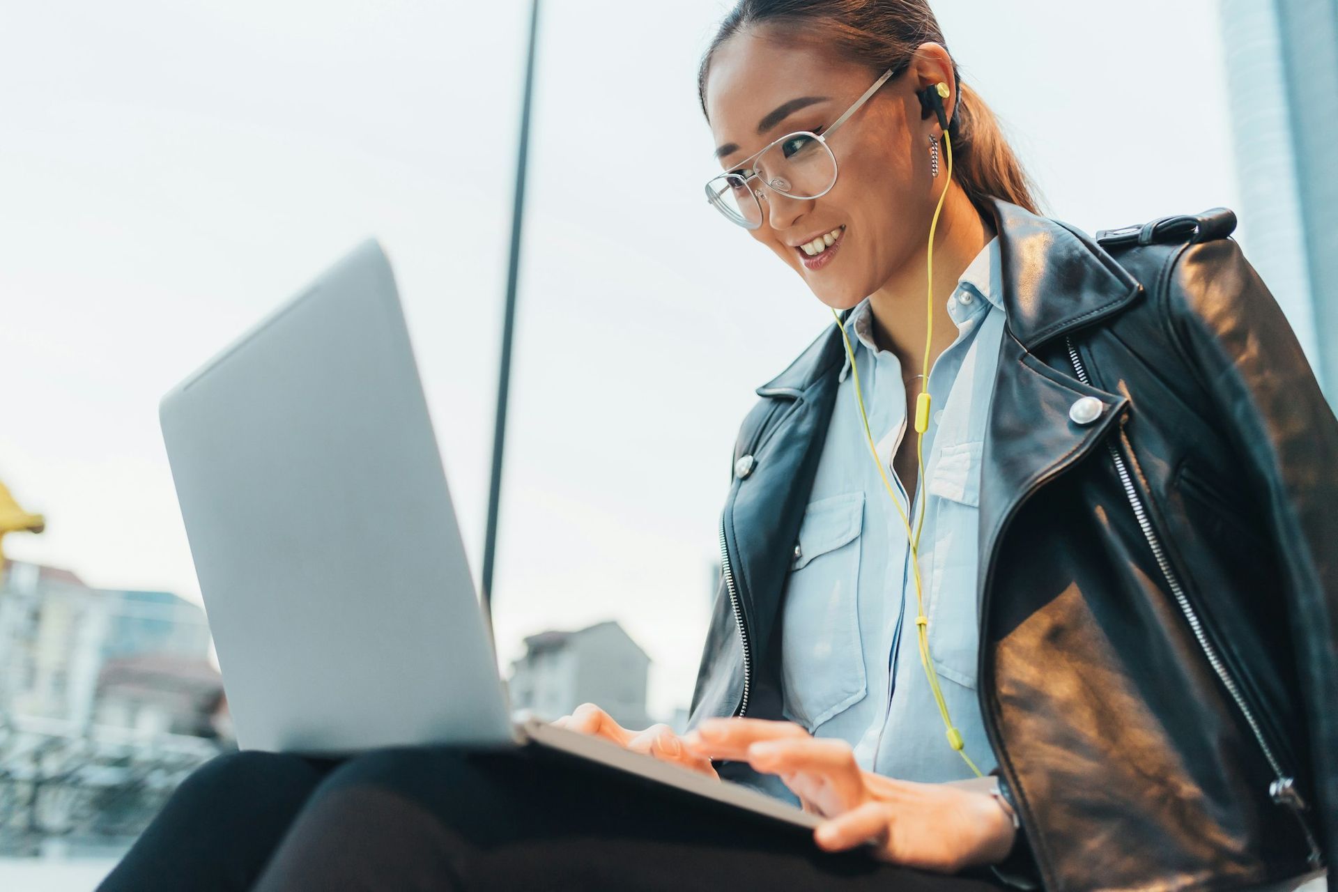 Woman with glasses and a black leather jacket works on a laptop outdoors.