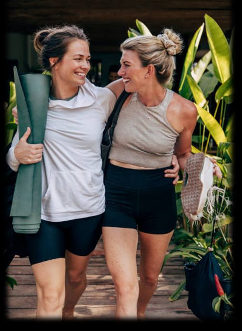 Two women smiling, arm in arm. One holds a yoga mat, the other a pair of shoes. Tropical setting. Two women smiling, arm in arm. One holds a yoga mat, the other a pair of shoes. Tropical setting.