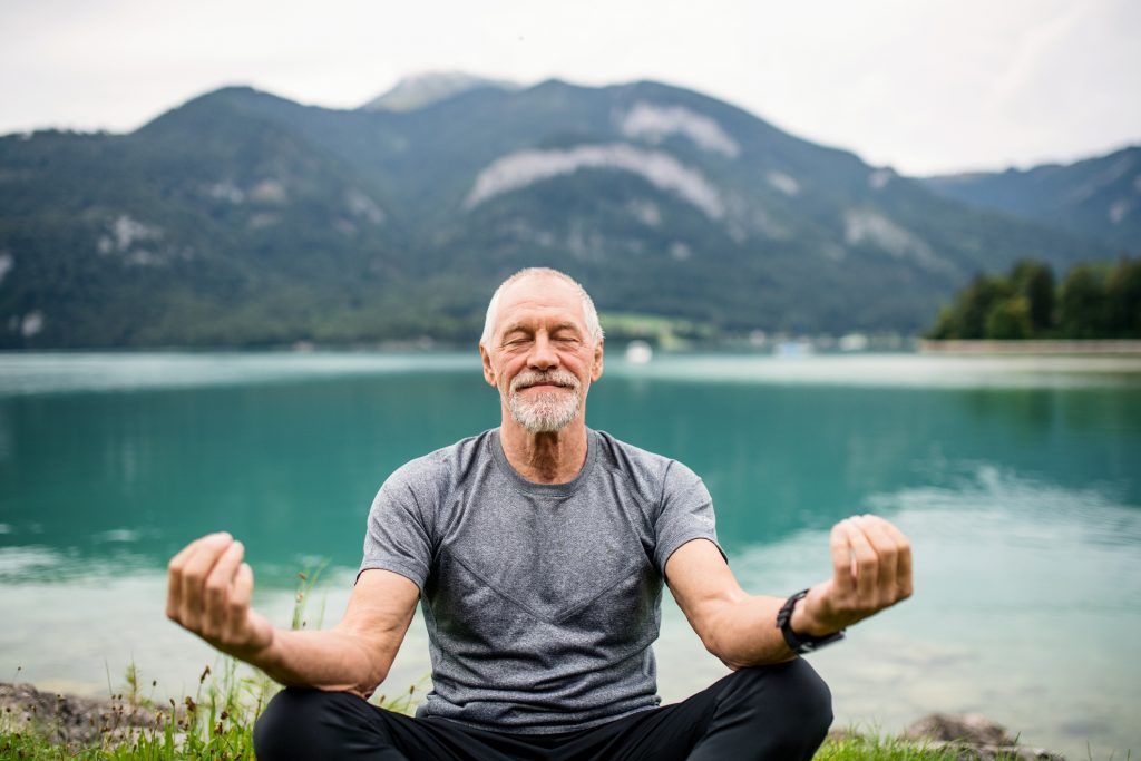 um homem mais velho está sentado em posição de lótus à beira de um lago com montanhas ao fundo.