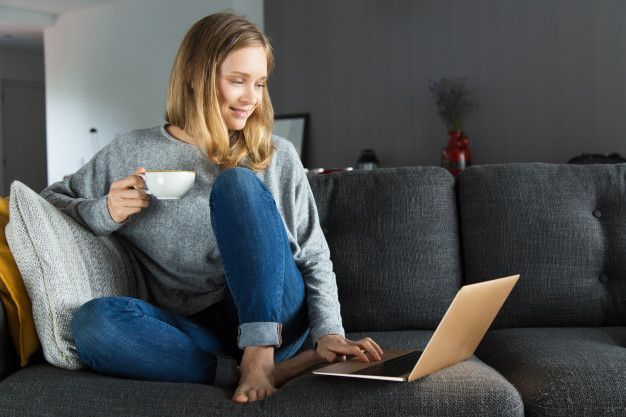 uma mulher está sentada em um sofá usando um laptop e tomando café.