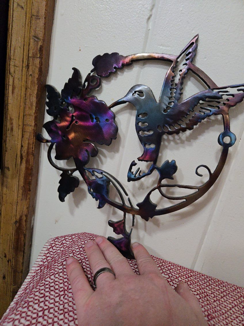 A person is holding a metal hummingbird and flower sculpture on a wall.