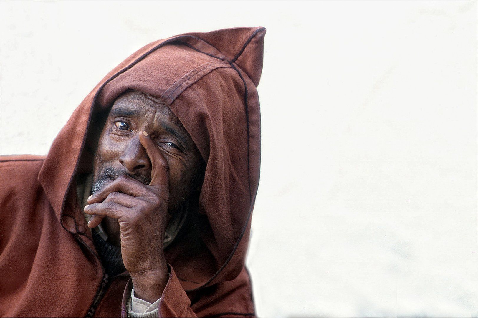A man wearing a brown hooded cape is covering his face with his hand.