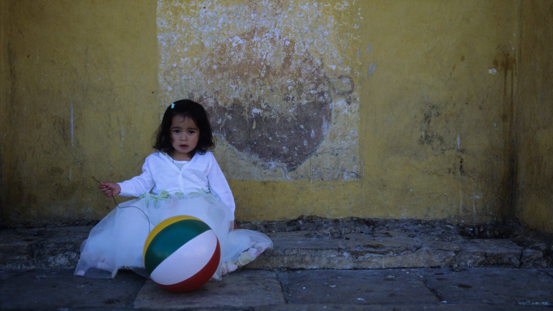 A little girl in a white dress is sitting on the ground with a beach ball.