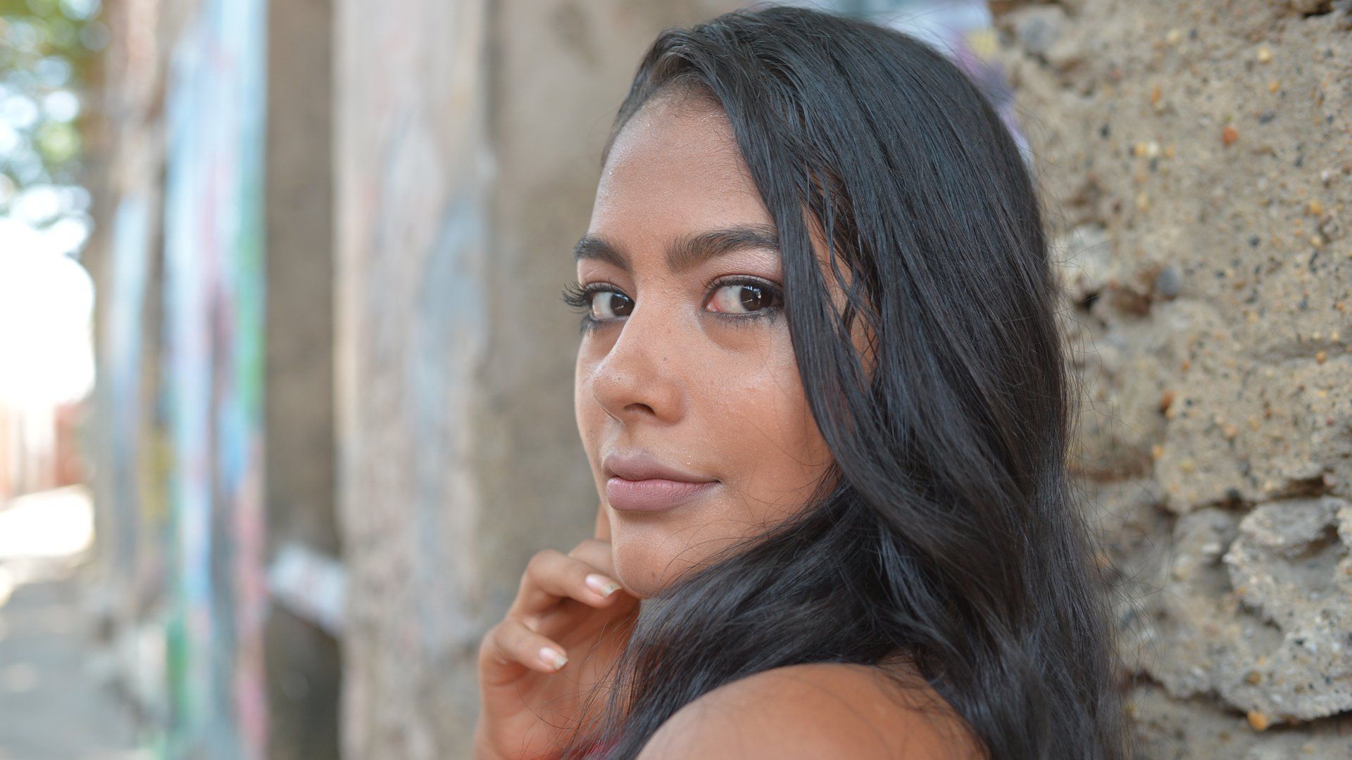 A woman with long black hair is leaning against a stone wall.