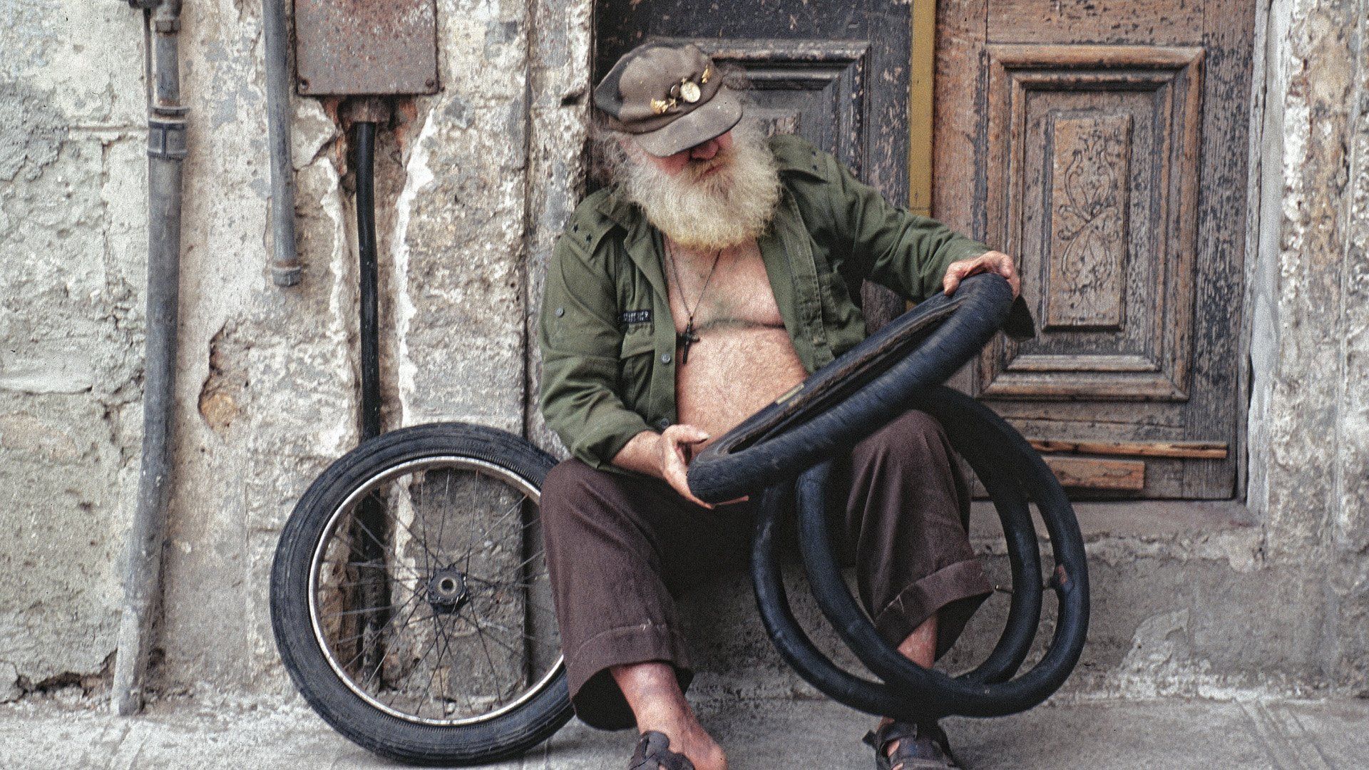 A man with a beard is sitting on the sidewalk holding a tire