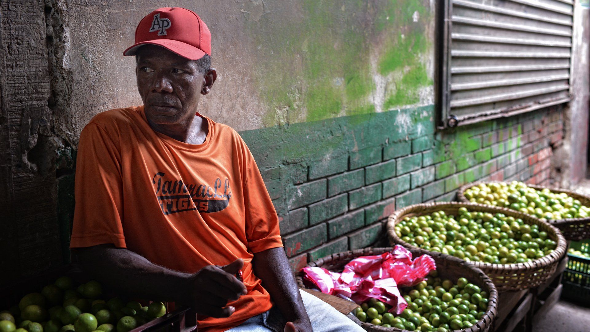 A man is sitting in front of a brick wall surrounded by baskets of fruit.