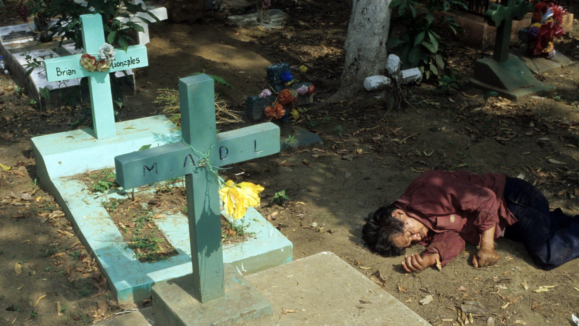A person is laying on the ground in front of a cross in a cemetery.