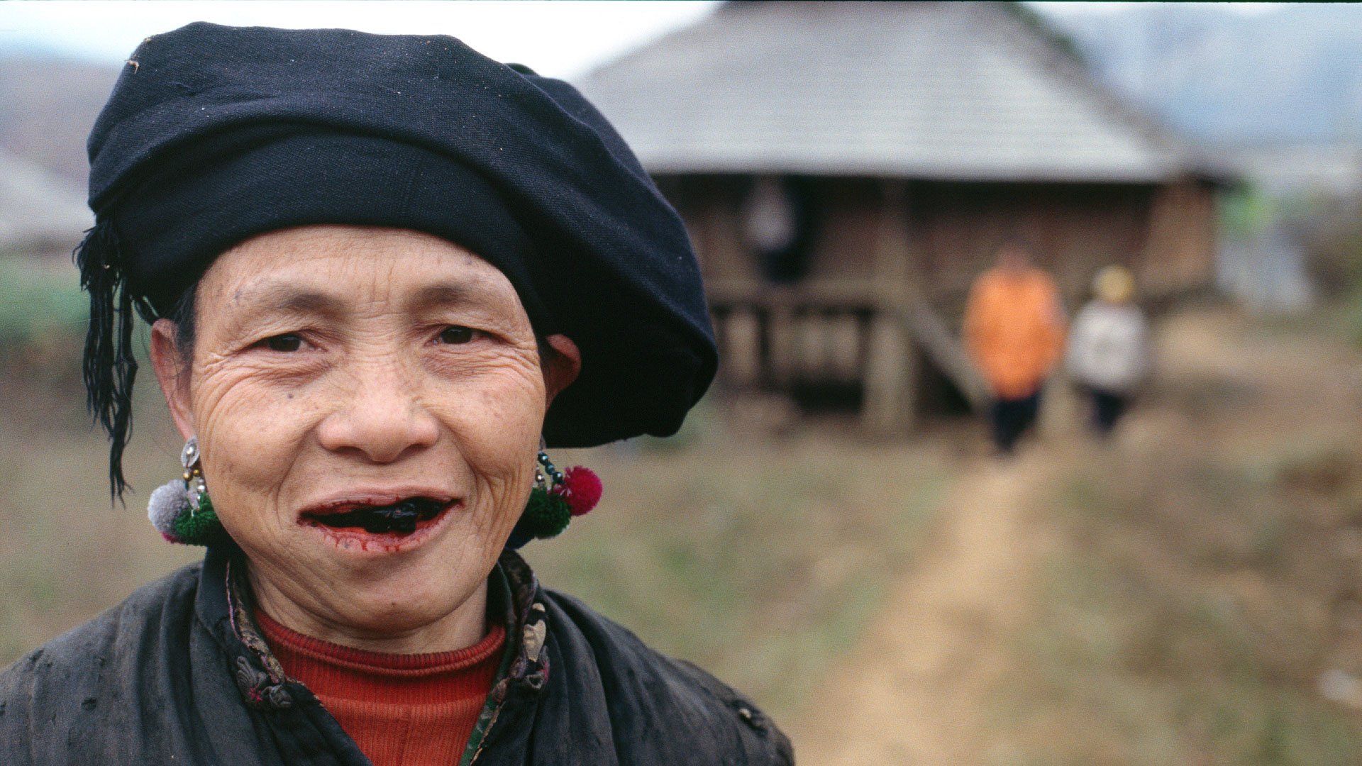 A woman with a black hat and earrings is smiling in front of a house.