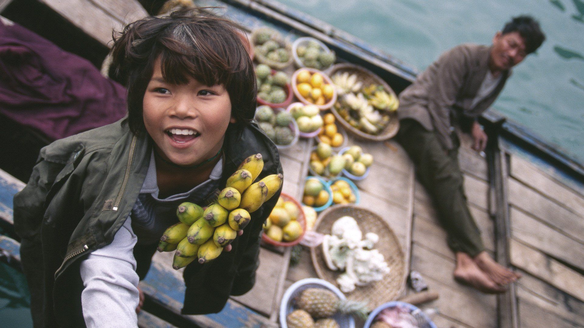 A young girl is standing on a boat holding a bunch of bananas.